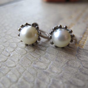 May include: A pair of vintage pearl earrings. Each earring features a large, round, creamy white pearl surrounded by a silver-toned, beaded frame. The earrings have a screw-back closure. The background is a textured, light-colored surface.