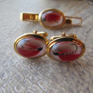 Peut inclure: Un ensemble de boutons de manchette et d'attache-cravate dorés. Chaque pièce présente un design ovale avec un couvercle transparent en forme de dôme mettant en valeur une mouche de pêche rouge et noire. L'ensemble a un style classique et élégant.