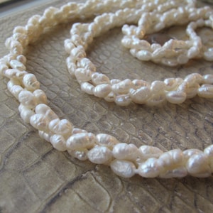May include: A close-up shot of a cream-colored pearl necklace. The necklace is composed of multiple strands of small, irregularly shaped pearls. The pearls have a subtle luster and are arranged in a twisted pattern. The background is a textured, neutral surface.