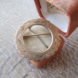 May include: A gold-toned circular brooch with a scalloped edge and leaf design, resting in an open, vintage, peach-colored jewelry box. The brooch features a pin closure and a central open circle.