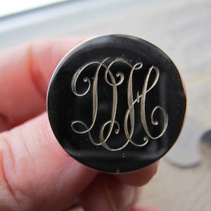 Op de afbeelding: Een rond, zwart metalen object met een zilveren monogram van de letters "JIH" in een elegant, cursief lettertype. Het object wordt in een hand gehouden, met een vage achtergrond.