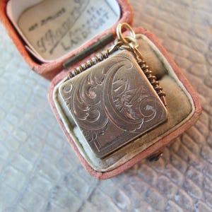 May include: A vintage square locket with intricate floral engravings, suspended from a delicate chain. The locket is displayed open within a pink velvet jewelry box, showcasing its detailed design and aged appearance.