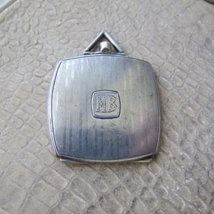 May include: A silver-toned compact with a rectangular shape and rounded corners. The compact features vertical line detailing and a small, square emblem with the letters "MB" in the center. A triangular top is attached.