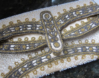 May include: A white beaded clutch bag with a gold and silver beaded design. The clasp is a large, ornate, gold-toned metal piece.