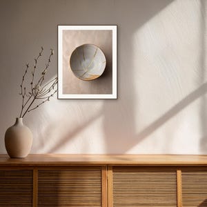 May include: A framed wall art print featuring a light-colored ceramic bowl with gold-colored cracks across its surface. The bowl is displayed in a room with a wooden cabinet, a vase with bare branches, and sunlight casting shadows on the wall.