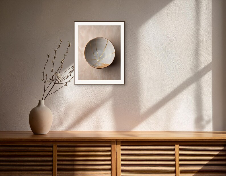May include: A framed wall art print featuring a light-colored ceramic bowl with gold-colored cracks across its surface. The bowl is displayed in a room with a wooden cabinet, a vase with bare branches, and sunlight casting shadows on the wall.