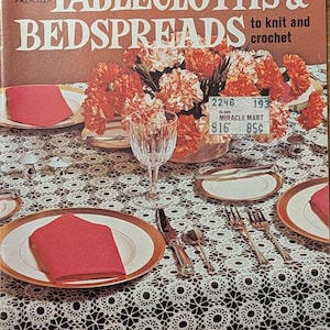 May include: A vintage Coats & Clark's Book No. 193 titled Tablecloths & Bedspreads to knit and crochet. The cover features a photograph of a dining table set with a white lace tablecloth, red napkins, plates, silverware, and orange flowers.