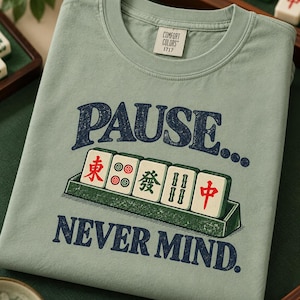 Könnte beinhalten: Ein salbeigrünes T-Shirt mit einer Grafik von fünf Mahjong-Steinen in einem Halter. Der Text darüber lautet PAUSE... und der Text darunter lautet NEVER MIND. Das Etikett Comfort Colors 1717 ist am inneren Halsausschnitt sichtbar.