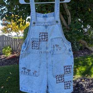 May include: Light blue denim overalls featuring brown screen-printed designs. The front displays two square quilt-style patterns on the chest and lower leg, alongside illustrations of various mushrooms on the chest pocket and lower left leg.