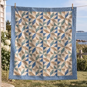 Puede incluir: Una colcha cuadrada con un patrón geométrico repetitivo de triángulos en tonos beige, azul claro y azul oscuro. La colcha está enmarcada por un borde azul con un pequeño estampado floral y está colgada de una cuerda con pinzas de madera.