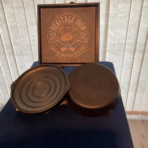 May include: Two black cast iron skillets are displayed in front of a framed sign. The sign features the text "HERITAGE IRON" and "CRAFTED ONE PIECE AT A TIME" with an illustration of a skillet and two small American flags in a circular design.