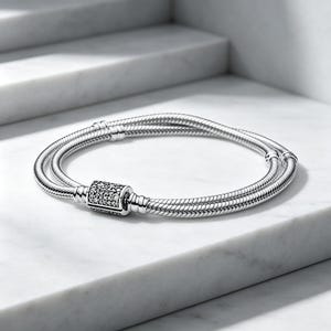 May include: A silver snake chain bracelet featuring a double-strand design and a cylindrical clasp encrusted with small, clear, sparkling stones. The bracelet has a polished metallic finish and a secure, integrated locking mechanism.