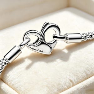 Könnte beinhalten: Ein silbernes Schlangenkettenarmband mit einem herzförmigen Verschluss und einem Unendlichkeitssymbol. Der Verschluss ist mit dem Wort PANDORA graviert. Das Armband hat eine strukturierte, flexible Gliederkette und eine polierte, glänzende Oberfläche.