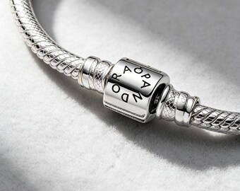 May include: A silver snake chain bracelet featuring a polished barrel-shaped clasp. The clasp is engraved with the brand name PANDORA in a circular pattern. The bracelet has a textured, woven metal design throughout the entire length of the band.