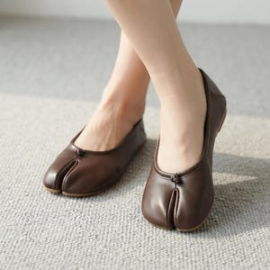 May include: A pair of dark brown leather tabi shoes featuring a split-toe design and a small decorative knot detail at the center of the vamp. These slip-on shoes have a low-cut topline and a smooth, matte finish, providing a clean look.