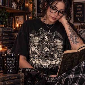May include: A black t-shirt featuring a graphic illustration of a skeleton wearing a witch hat and reading a book, surrounded by stacks of books, a raven, and a cat. The text on the shirt reads THE BOOKWORM, and the shirt is worn with black glasses.