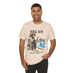 Puede incluir: Una camiseta de manga corta color beige claro con un gráfico de una cabra con sombrero y peto, sosteniendo una paleta de albañil. El texto dice SOY UN PARA LAS ALBERCAS. El gráfico incluye una piscina, herramientas y cubos.