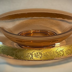 May include: A round, peach-colored glass bowl featuring a wide, flat rim decorated with a gold-toned, etched floral pattern. The center of the bowl is recessed and circular, with the overall design showcasing a translucent, smooth glass finish.