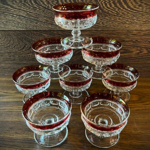 May include: A set of eight clear glass dessert cups arranged in a pyramid formation. Each cup features a deep red rim and a textured, pressed glass pattern on the bowl and base. The stems are short and sturdy, supporting the wide, circular bowls.