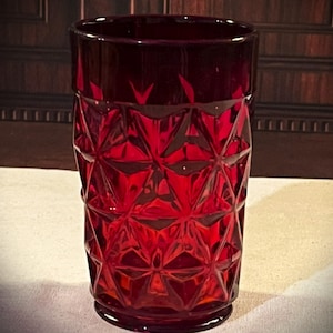 May include: A deep red glass tumbler featuring a prominent geometric pattern of raised triangular facets across its exterior surface. The vessel has a cylindrical shape with a smooth rim and a circular base, showcasing a translucent ruby hue.