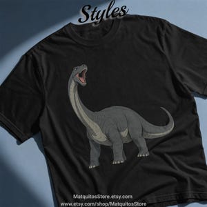 Könnte beinhalten: Ein schwarzes T-Shirt mit kurzen Ärmeln und einer Grafik eines grauen Langhals-Dinosauriers mit offenem Maul. Das Wort Styles ist oben in Schreibschrift gedruckt, und der Text MatquitosStore.etsy.com steht am unteren Rand.