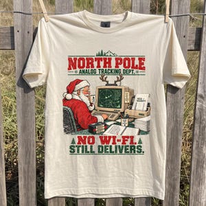 May include: A cream-colored t-shirt featuring a graphic of Santa Claus at a computer with the text NORTH POLE ANALOG TRACKING DEPT. and NO WI-FI. STILL DELIVERS. The design includes a radar screen, a printer, and a checklist on a desk.