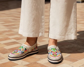 May include: A pair of slip-on shoes featuring intricate, multi-colored geometric embroidery in shades of pink, green, blue, and yellow on a cream-colored base, set on a flat, layered sole with a black trim and wide-leg cream trousers.