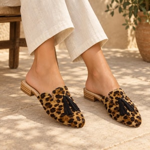 May include: A pair of slip-on mules featuring a brown and black leopard print pattern. Each shoe has a pointed toe, a small stacked wooden heel, and a decorative black tassel centered on the top of the vamp. The material has a textured finish.