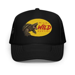 Puede incluir: Una gorra tipo trucker negra con un parche ovalado amarillo en la parte frontal. El parche muestra una ilustración detallada de una tortuga mordedora con la boca abierta y el texto rojo MIAMI WILD EXOTICS en letra negrita.