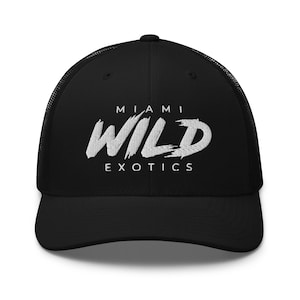 May include: A black trucker cap featuring a mesh back and a solid front panel. The front displays white embroidered text that reads MIAMI WILD EXOTICS, with the word WILD in a large, stylised, brush-stroke font and MIAMI and EXOTICS in smaller text.