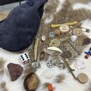 May include: A collection of mystical items including a dark blue suede pouch, dice, wooden runes, a seashell, a small sword, and other charms. The items are arranged on a cowhide rug.