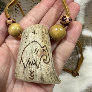 May include: A handcrafted necklace featuring a pendant carved with an elephant design and a cross. The pendant is made of a light-colored material, possibly bone or wood, and is strung on a leather cord with wooden beads.