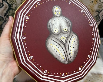 May include: A maroon, octagonal decorative plaque featuring a central hand-painted figure of a stylized, curvaceous person in shades of gray with yellow and white dotted accents. The border is decorated with white geometric lines and yellow dots.