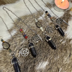 May include: A selection of crystal pendulums with silver chains and decorative charms. The pendulums feature dark blue crystals, pentagrams, feathers, and beaded accents. A lit candle is in the background.