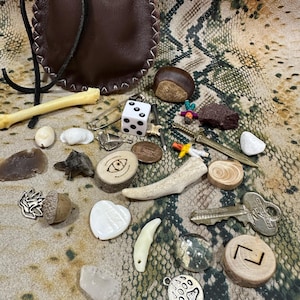 May include: A dark brown leather drawstring pouch accompanied by a collection of small objects including a white die, a metal key, a miniature sword, a wooden slice with an eye symbol, a small bone, a shell, a stone, and a metal bear paw charm.