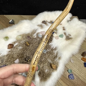 May include: A light brown wooden wand with carved symbols and a sun design. The wand is held against a backdrop of a white and brown fur-like material, with colorful gemstones scattered around.