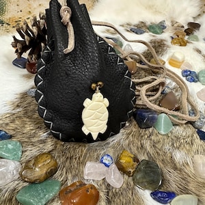 May include: A black leather pouch with a white turtle pendant. The pouch is decorated with white stitching and a drawstring closure. Various colorful gemstones are scattered around the pouch, including green, brown, and blue stones.