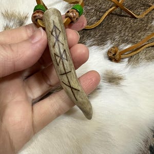 May include: A light brown antler pendant with etched symbols and a leather cord. The pendant has a cylindrical shape with a pointed end. Green and brown beads are attached to the leather cord near the top of the pendant.