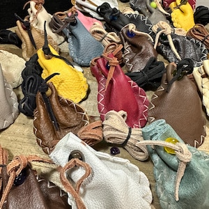 May include: A collection of small, multi-coloured leather drawstring pouches featuring visible whip-stitched edges, leather cord closures, and decorative beads. The pouches are rendered in various colours including white, brown, red, yellow, and blue.