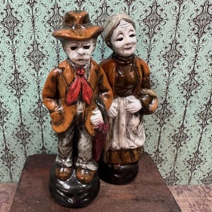 May include: A pair of vintage ceramic figurines depicting an elderly couple. The man wears a brown hat, coat, and red tie. The woman is in a brown dress and white apron. The figurines are on a dark wooden surface.