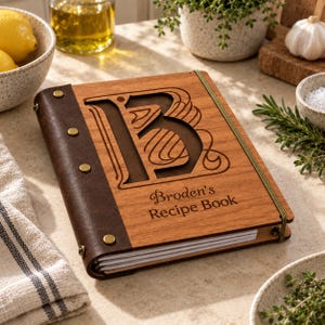 Op de afbeelding: Een rechthoekig receptenboek met een houten voorzijde voorzien van een grote lasergesneden letter B en de tekst Broden's Recipe Book. De kaft is bevestigd aan een donkerbruine lederen rug met vier messingkleurige metalen klinknagels en elastiek.