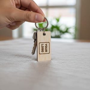 Op de afbeelding: Een rechthoekige lichtgekleurde houten sleutelhanger met een metalen ring en een metalen sleutel. Het houten oppervlak is gegraveerd met een vierkante rand en de tekst YOUR LOGO in het midden. De sleutelhanger heeft een gladde houtnerf.