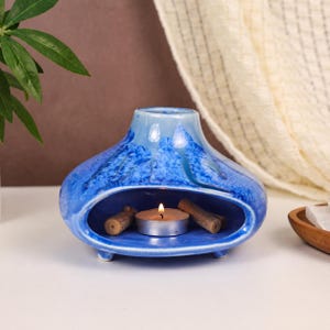 May include: A blue ceramic oil burner with a glossy, mottled glaze finish. The vessel features a wide, oval-shaped opening at the base containing a small, lit tea light candle and two small wooden sticks, with a narrow circular opening at the top.
