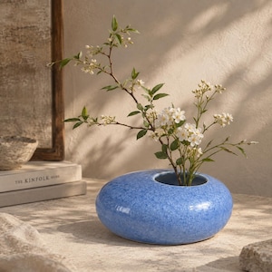 May include: A low, rounded ceramic vase with a vibrant blue mottled glaze and a smooth, pebble-like shape. The vase features a single circular opening at the top, currently holding a delicate branch with small green leaves and white flowers.