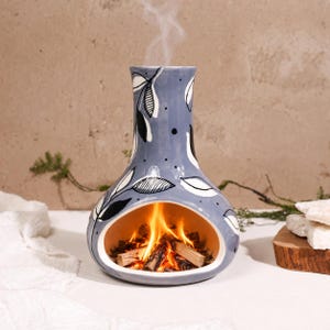 May include: A ceramic fireplace incense burner in a soft blue-grey hue, featuring a hand-painted black and white leaf pattern. The wide, oval-shaped opening contains burning wood pieces, emitting a thin trail of smoke from the top opening.