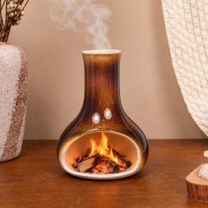 May include: A ceramic incense burner shaped like a miniature fireplace with a dark brown and tan gradient glaze. The base features an open hearth containing small wooden logs with orange flames and white smoke rising from the top chimney opening.