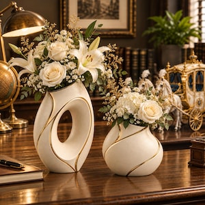 Pode incluir: Dois vasos de cerâmica em tom off-white com detalhes dourados, cada um com uma silhueta curva única. O vaso mais alto possui uma abertura oval central, enquanto o mais baixo é arredondado. Ambos contêm arranjos de rosas e lírios brancos.