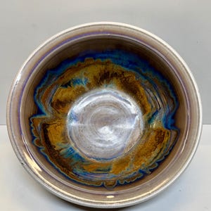 May include: A round ceramic bowl featuring a glossy interior glaze with swirling patterns of deep blue, golden brown, and metallic silver. The rim is a smooth, light beige, and the overall finish is reflective, showing a smooth, curved surface.