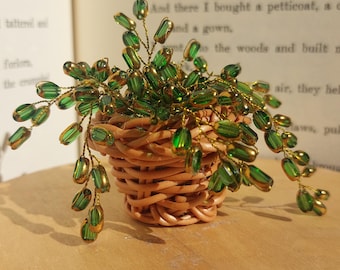 May include: A miniature decorative plant crafted from green glass beads on thin gold-toned wire, arranged in a small, round, light brown woven wicker basket. The plant features numerous translucent green bead leaves on flexible wire stems.