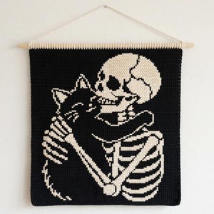 May include: A square crochet wall hanging featuring a black and off-white tapestry design of a human skeleton embracing a black cat. The piece is suspended from a light-colored wooden dowel with a thin white string attached for wall mounting.